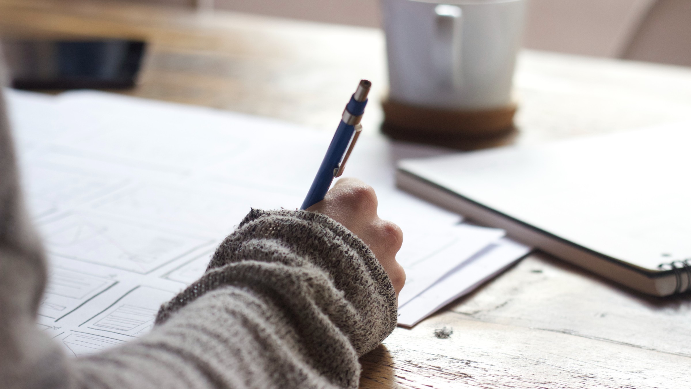 Someone writing at a wooden table next to a coffee mug